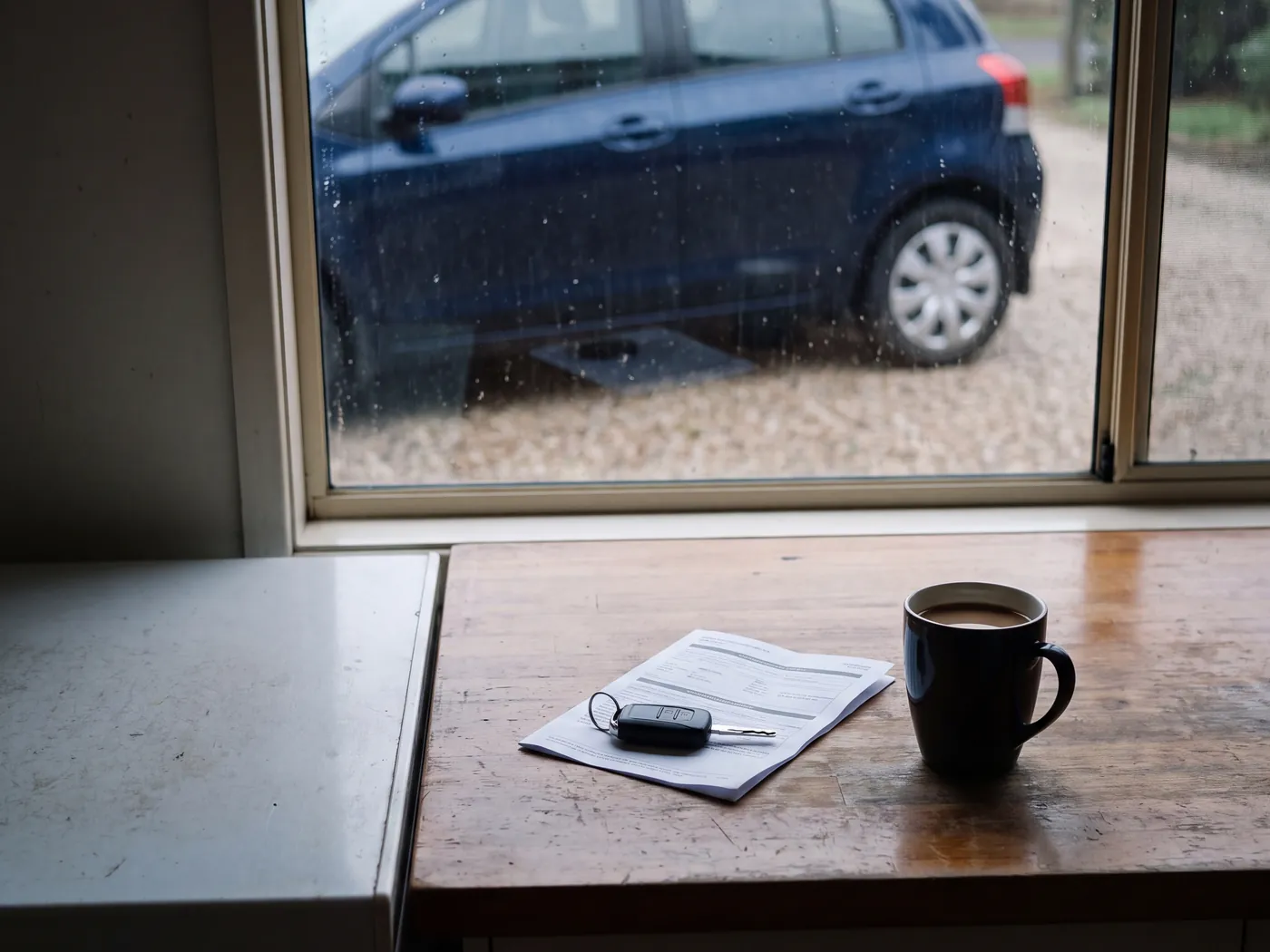Timber desk by a rain-flecked window with car keys, a loan document and a coffee, hatchback parked outside