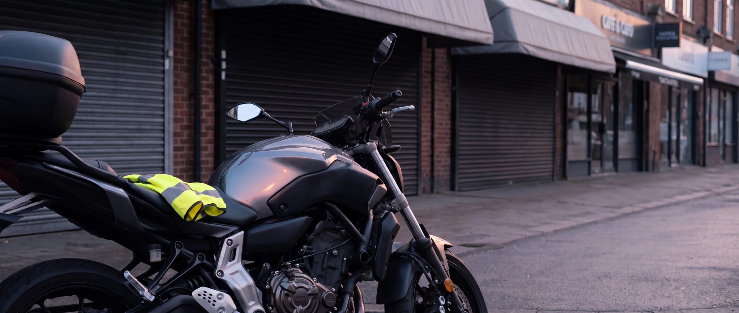 Grey motorbike with a top box and hi-vis vest on the seat outside early-morning shops