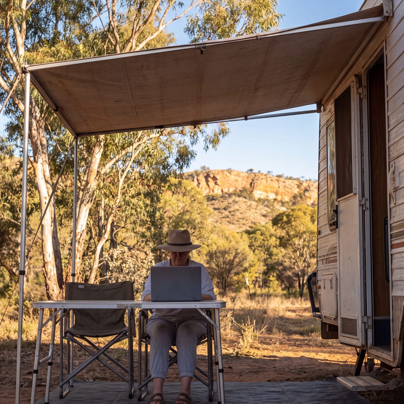 Person working on a laptop under a caravan awning at a bush campsite