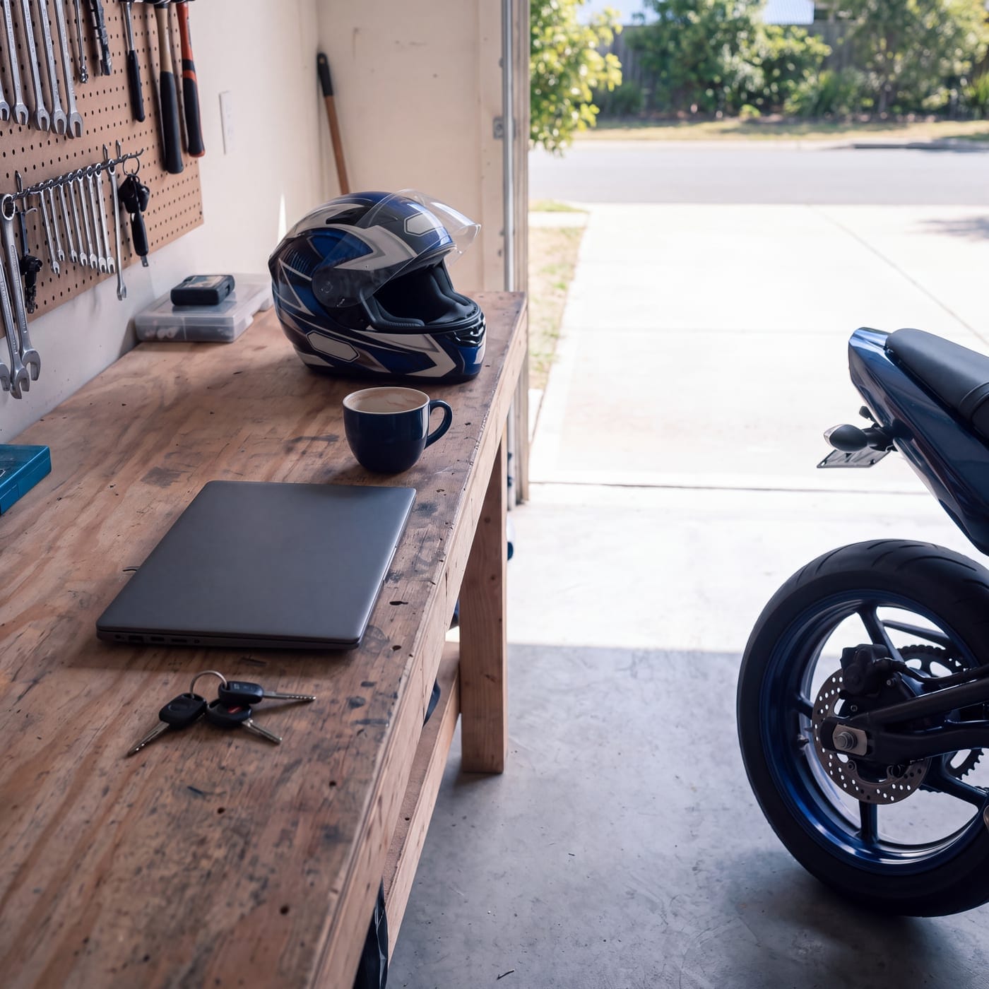 Garage workbench with a helmet, closed laptop, keys and a coffee, motorbike alongside