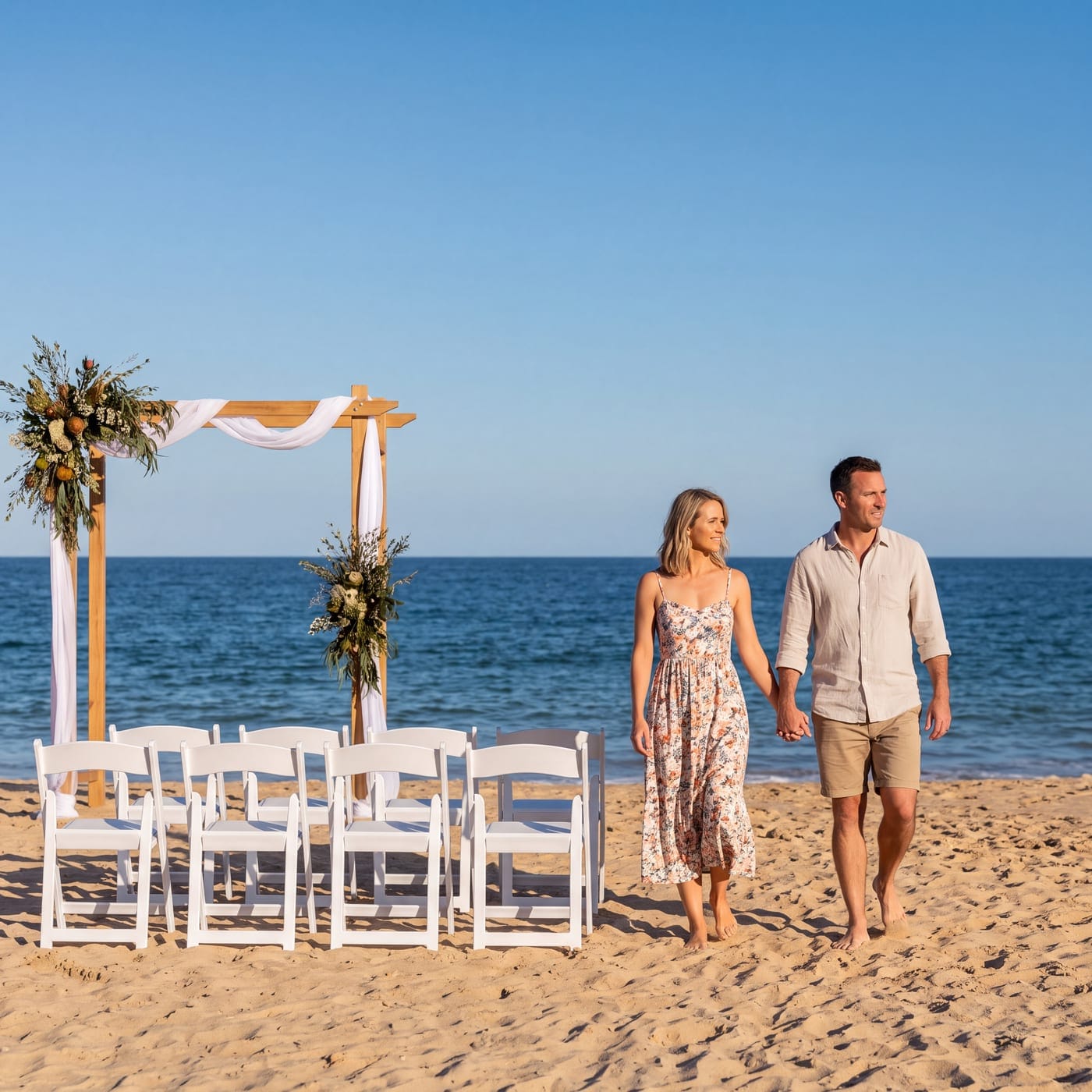 Couple walking hand in hand on a beach past a floral arbour and rows of white chairs